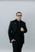 Man wearing a black suit and glasses against a plain background