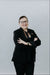 Person wearing a black suit and glasses with arms crossed against a plain background